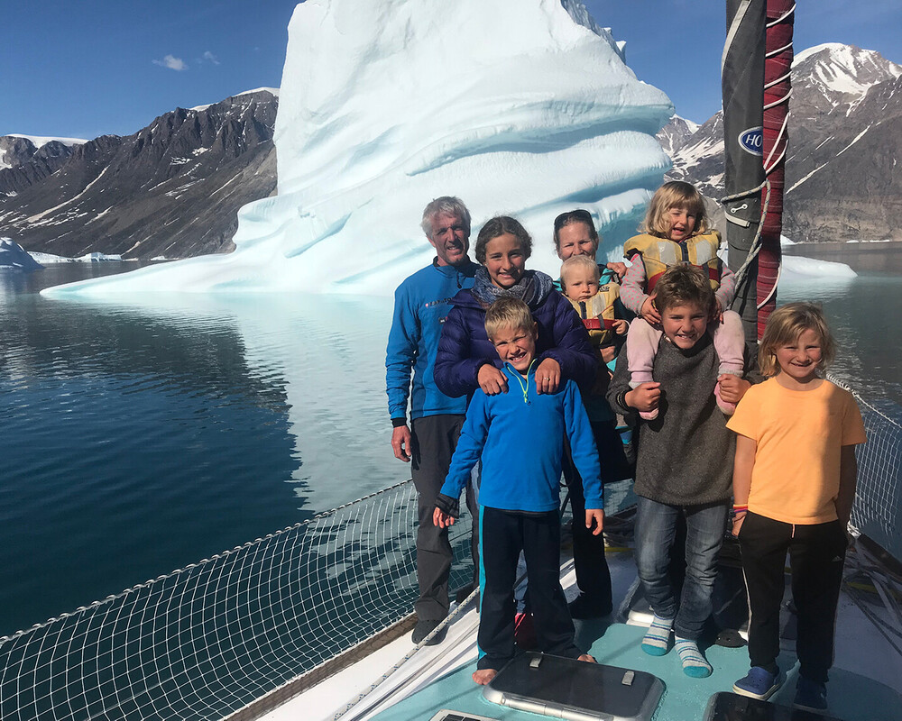The Schw&ouml;rer family posing together on the deck of their TOPtoTOP expedition sailboat in front of a towering iceberg in a Greenland fjord &ndash; parents and six children on a climate awareness voyage