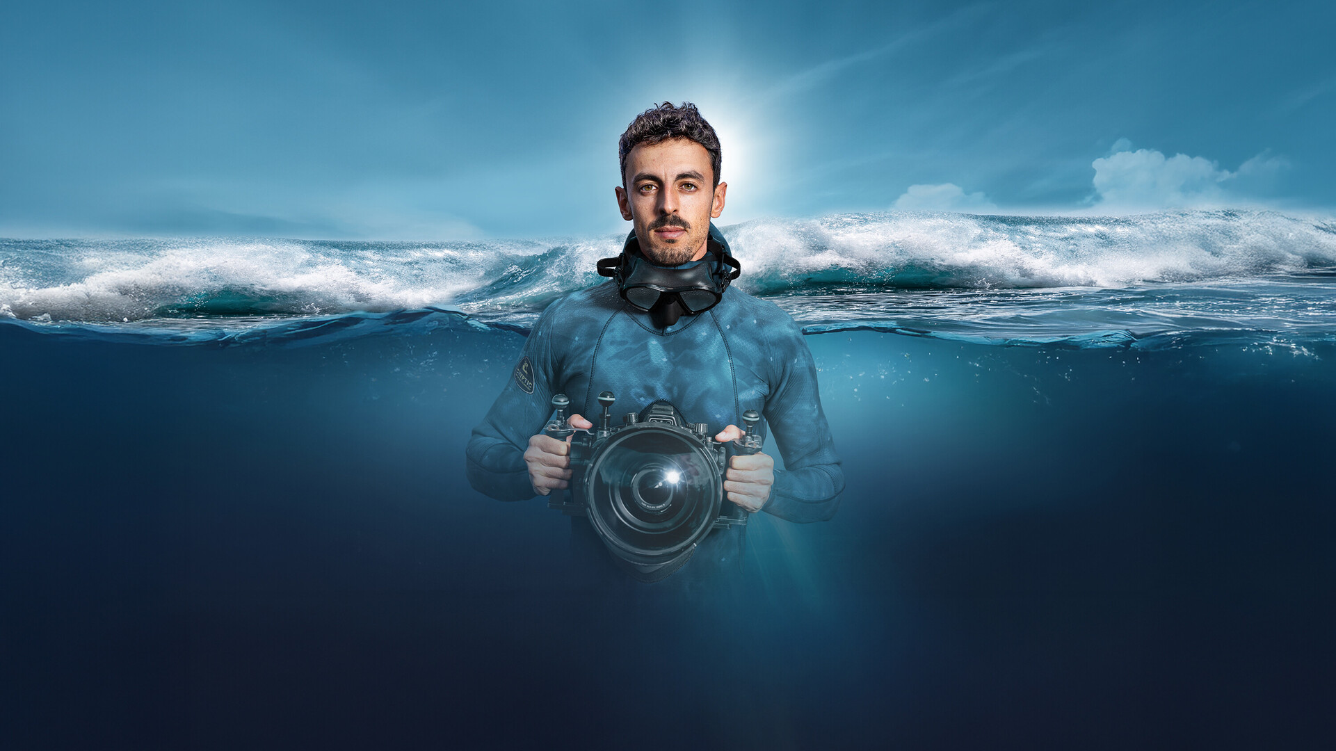 Plongeur avec masque et cam&eacute;ra sous-marine dans l'oc&eacute;an, &agrave; moiti&eacute; immerg&eacute;, avec une vague d&eacute;ferlante en arri&egrave;re-plan.